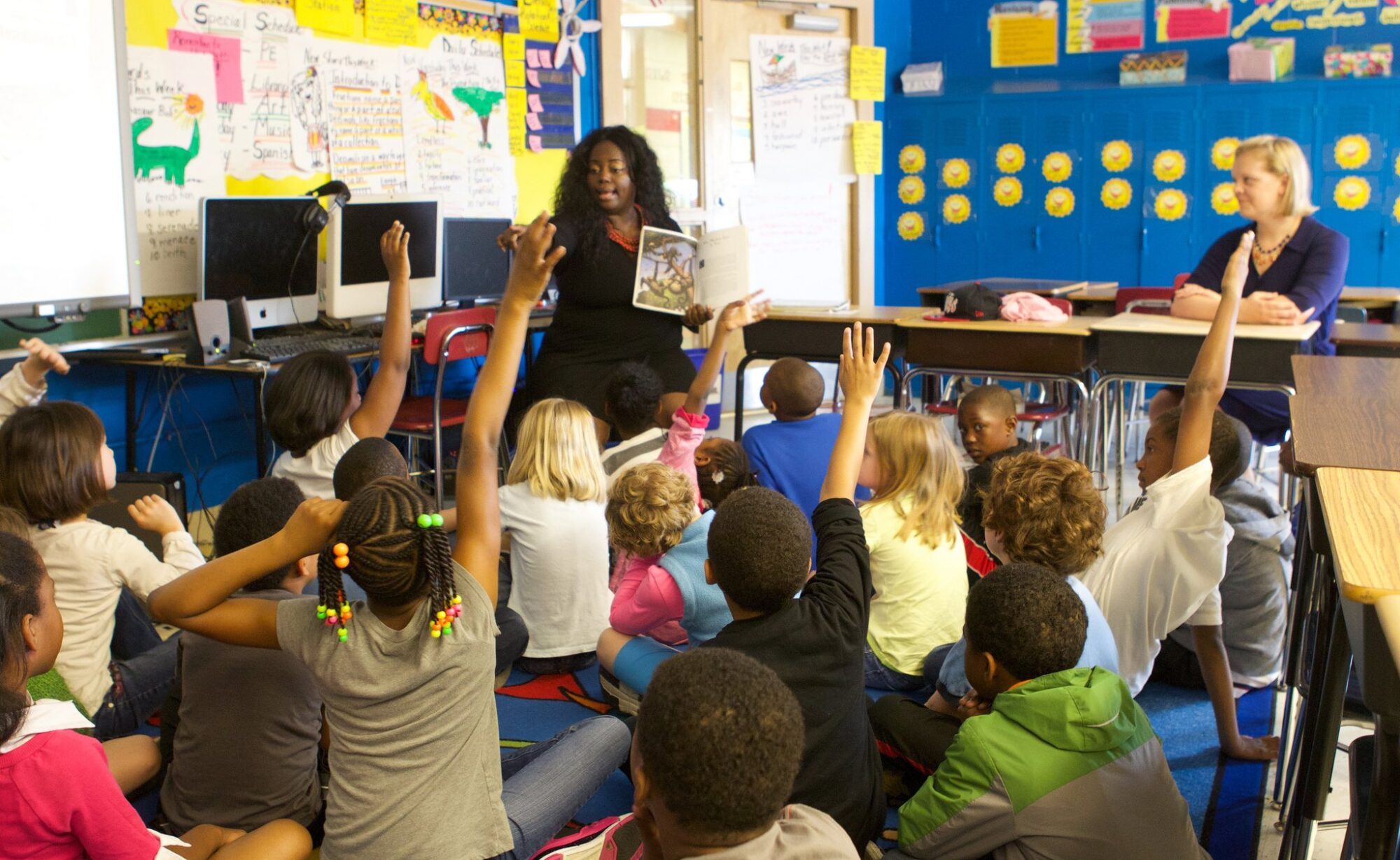 Children sitting on a classroom floor, raising hands, with a teacher reading a book aloud.