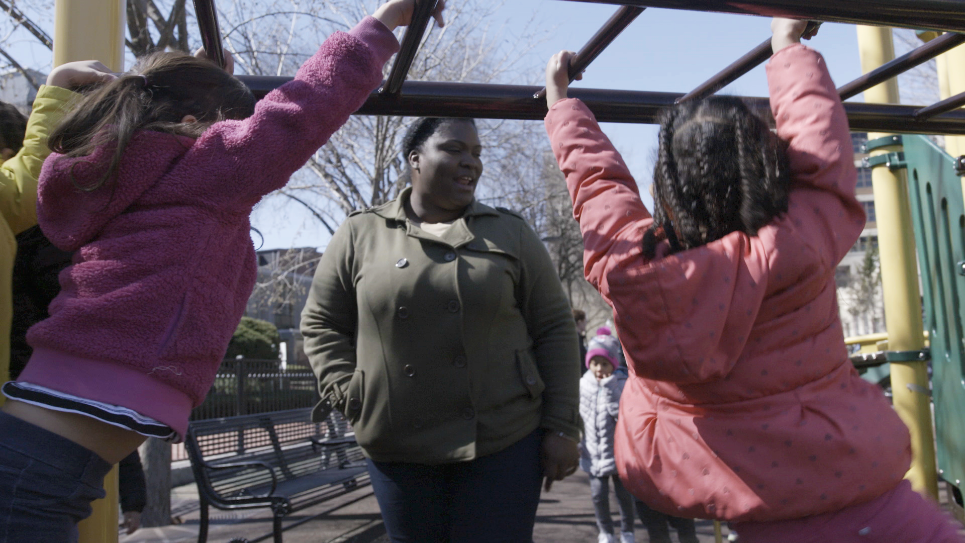 Two children on monkey bars with an adult watching in a playground setting.