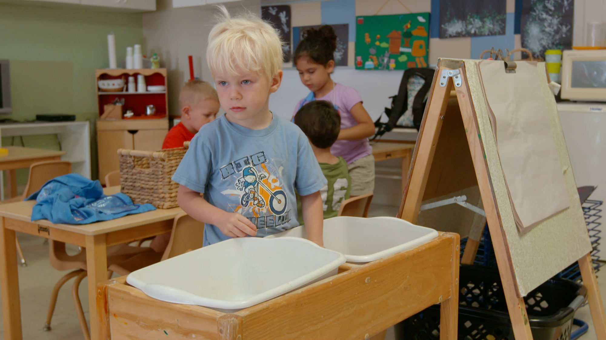 Children in a classroom setting with one child standing by a play station and others engaged in activities at a table.