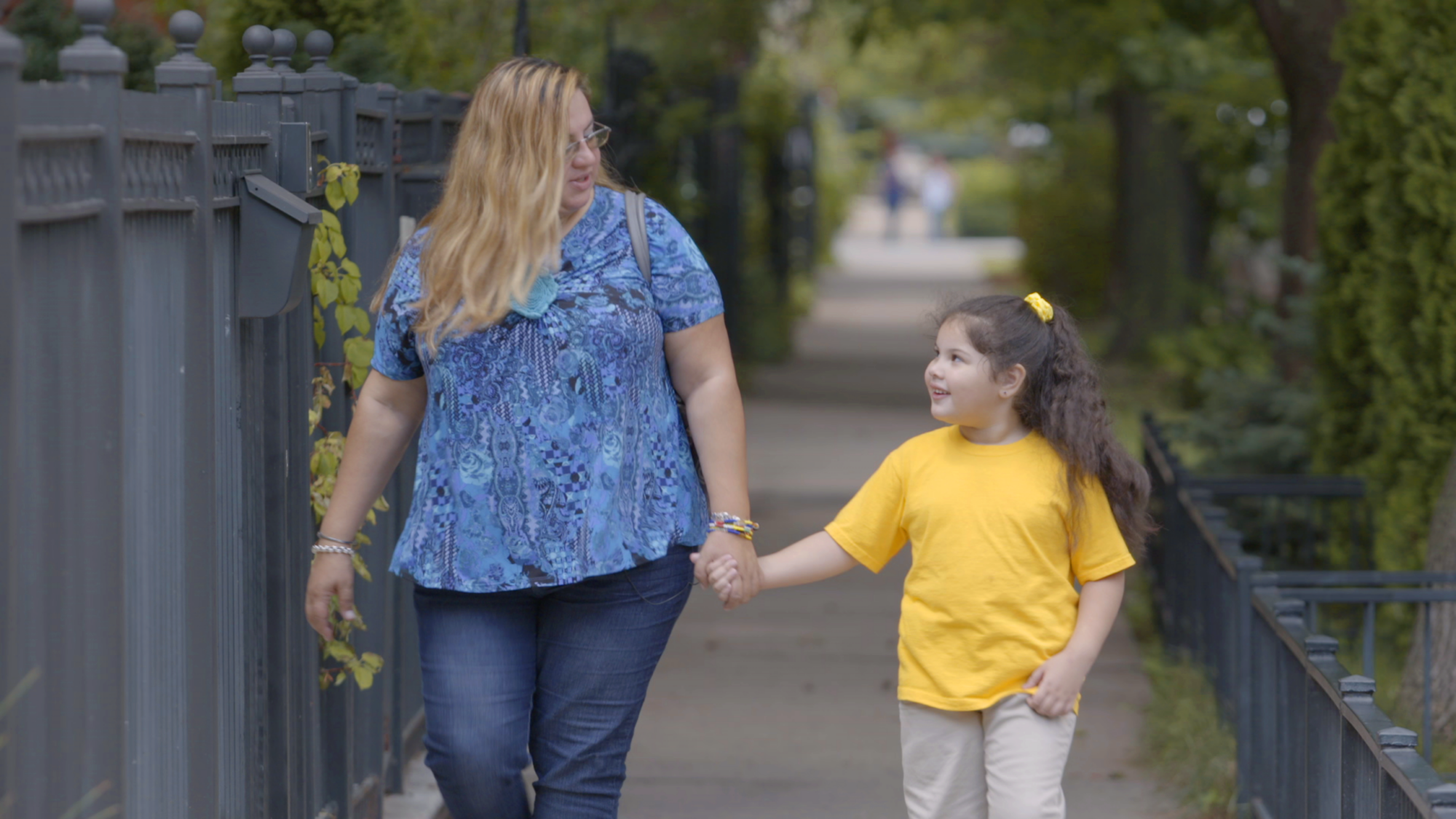 A woman and a young girl holding hands while walking on a sidewalk beside an iron fence.
