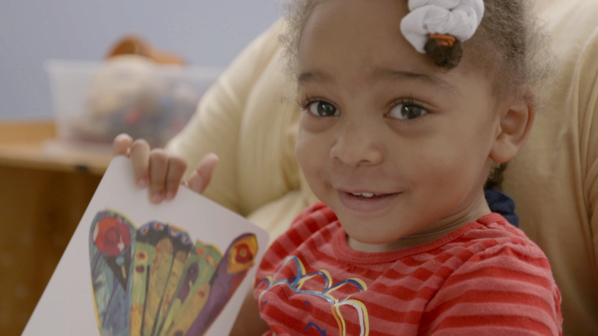 A smiling child in a red striped shirt holds a book with a colorful illustration.