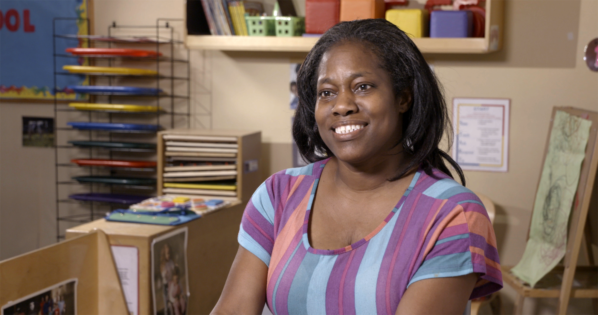 Woman smiling in a colorful shirt, seated in a classroom with art supplies in the background.