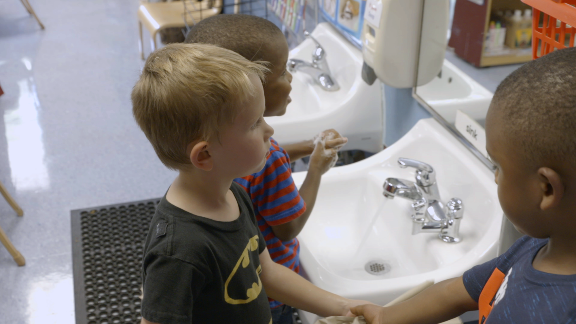 Three young boys washing hands at white sinks in a school restroom.