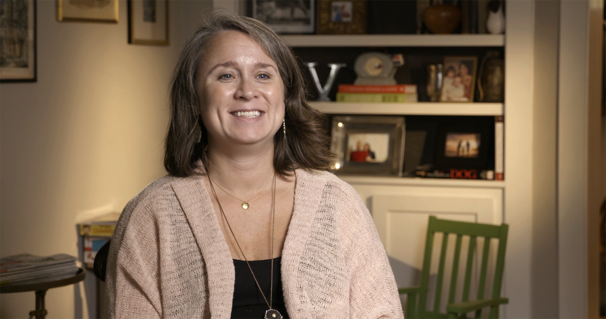 Person smiling in a room with shelves, framed photos, and decorative items in the background.