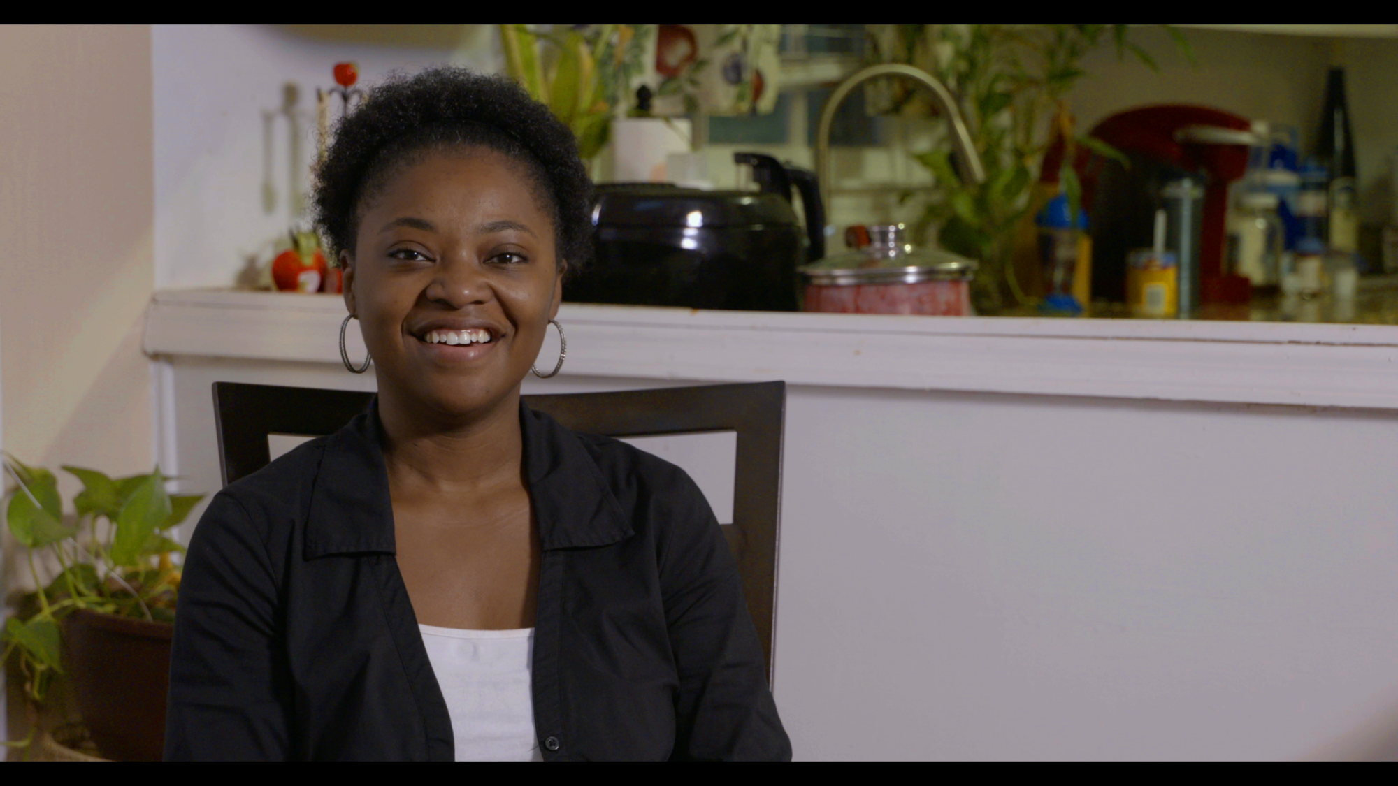 A smiling person sitting in front of a kitchen with a black pot and plants visible in the background.