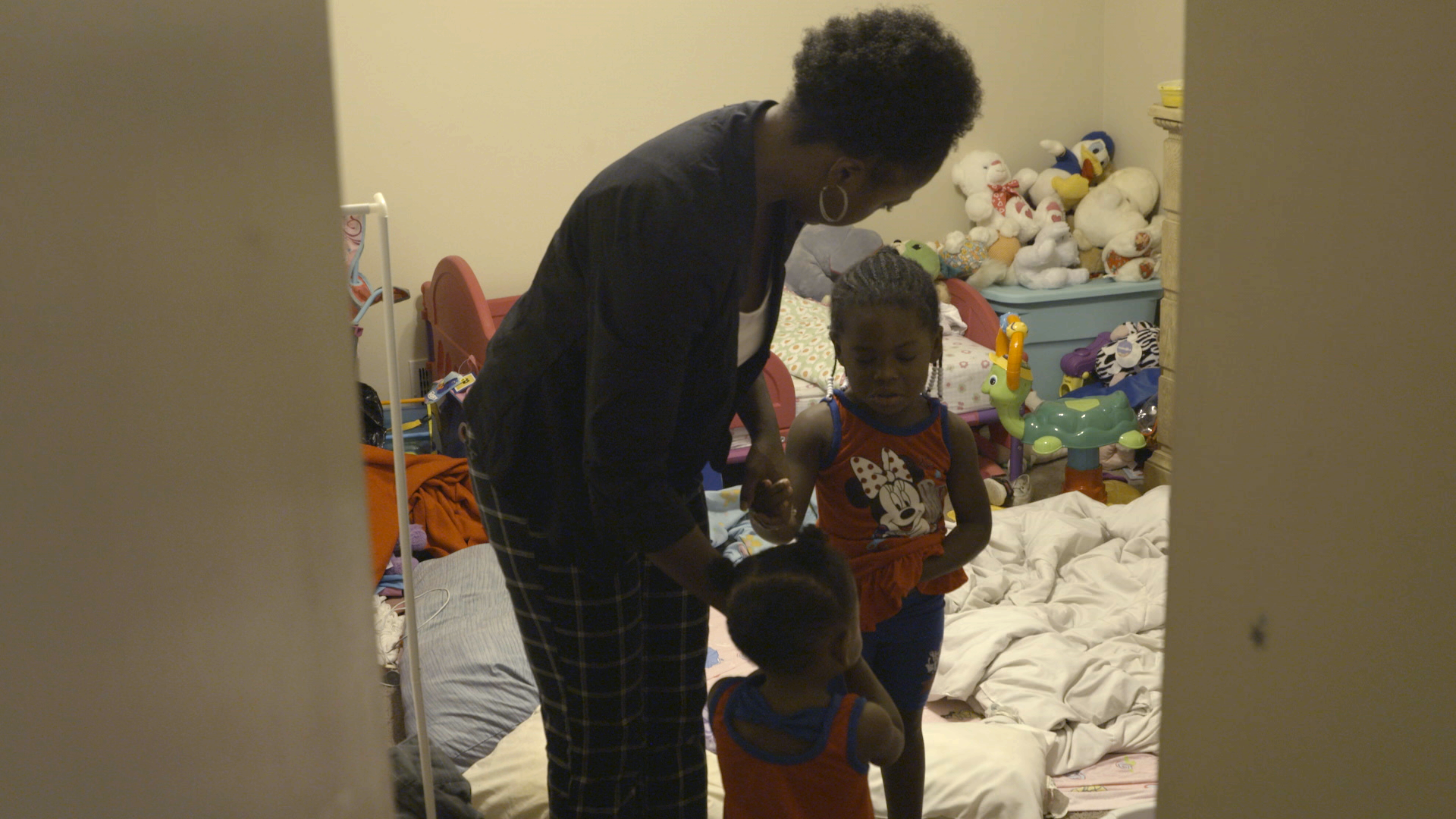 A woman bends down to interact with two young children in a toy-filled bedroom.
