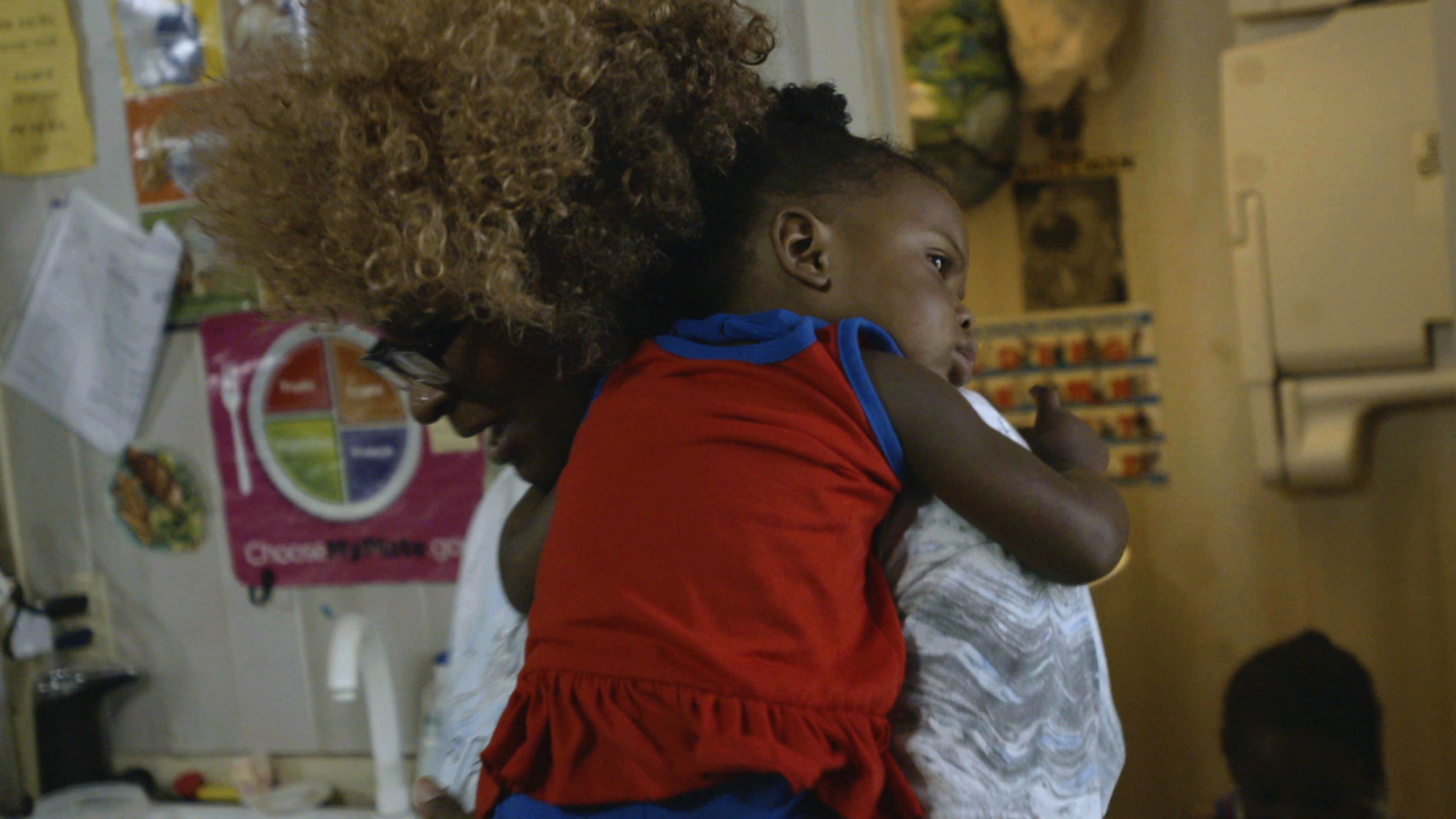 A woman holds a child clad in red with a kitchen setting in the background.
