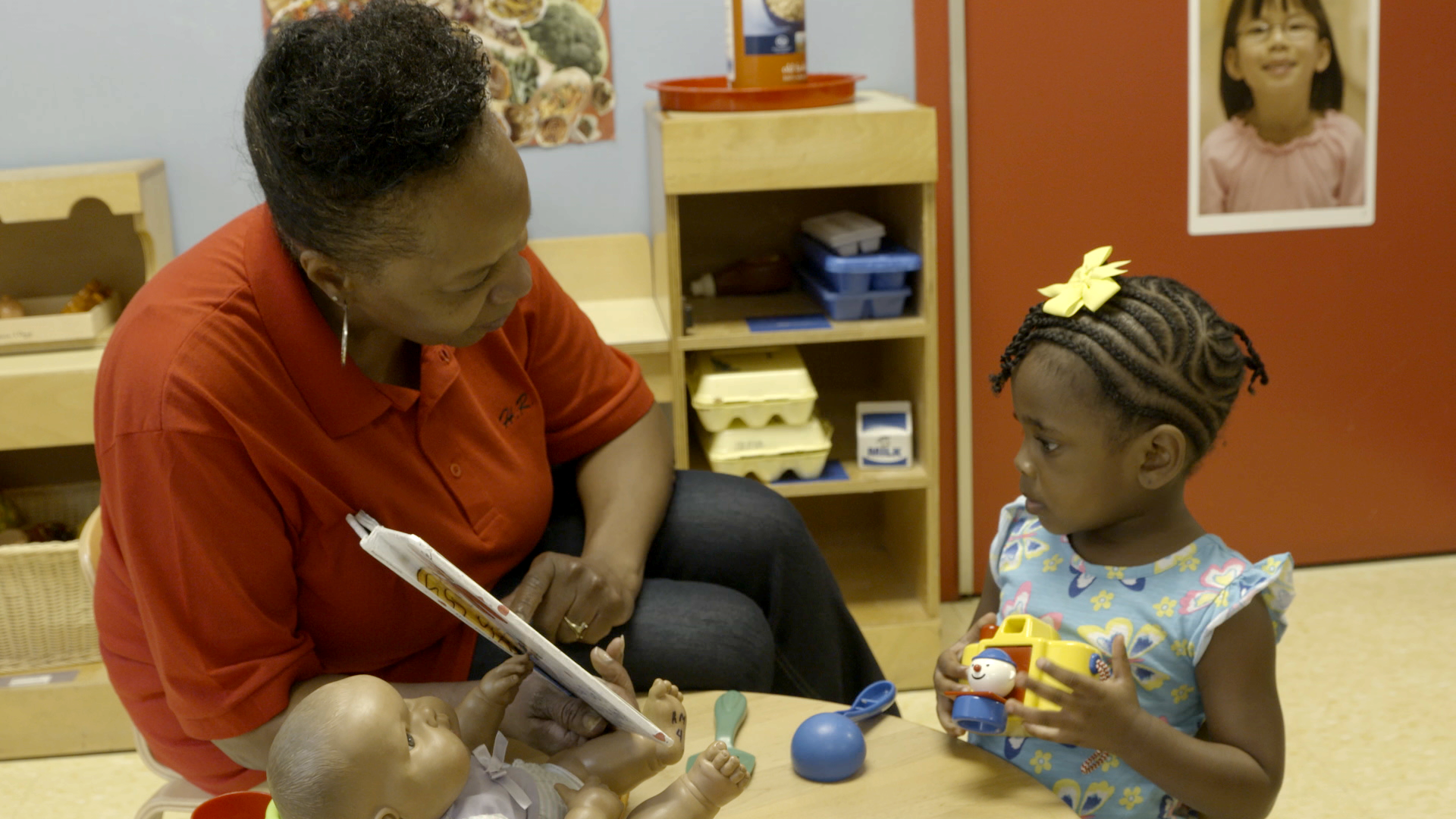 A woman in a red shirt reads to a young child holding a toy, with shelves and a poster in the background.