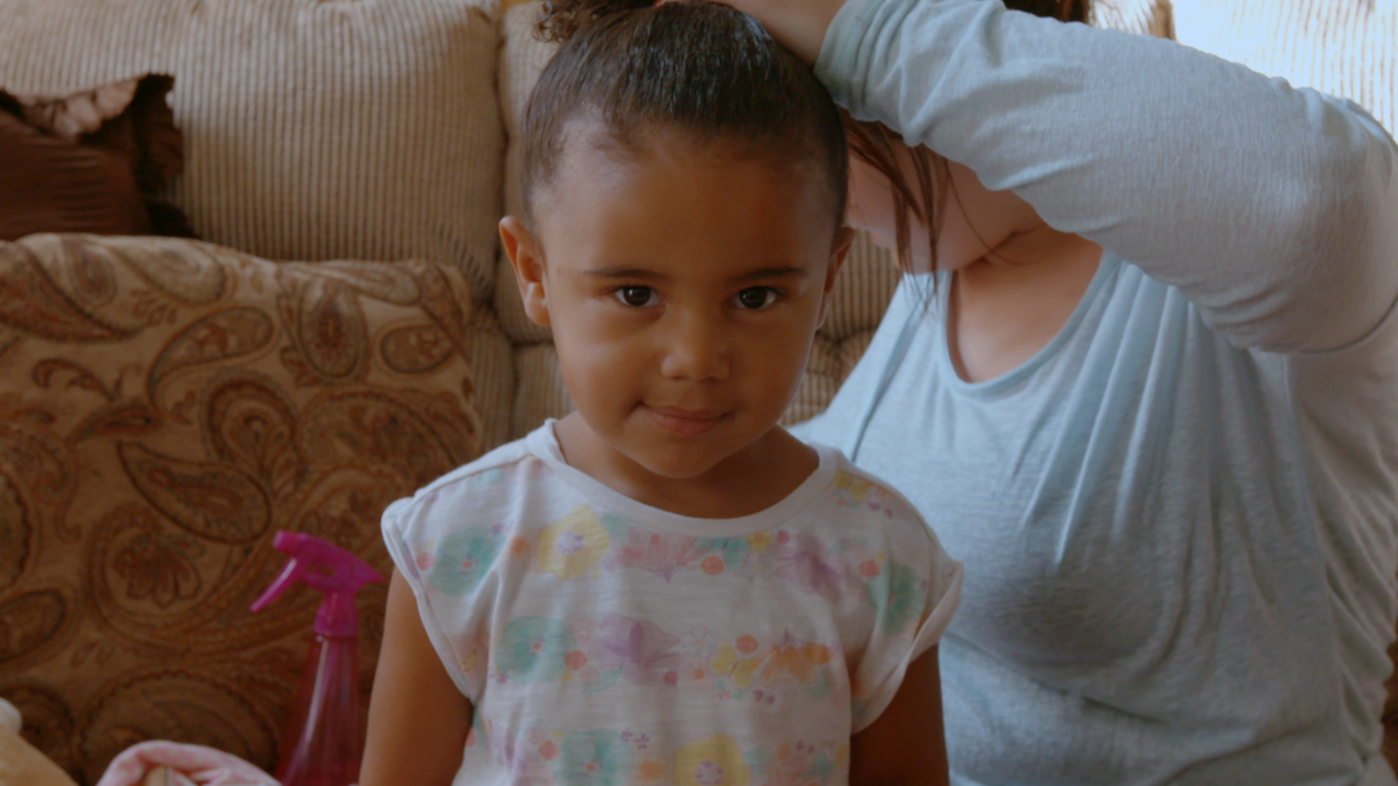 A young child with hair in a bun stands in front of an adult adjusting their hair on a couch.