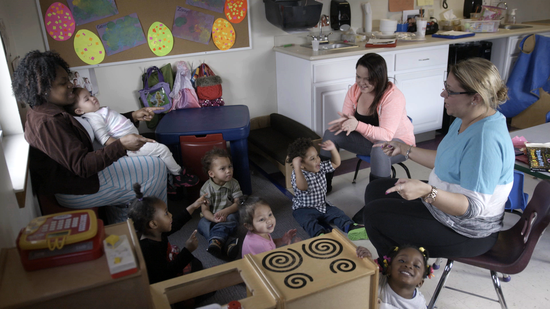 Children and caregivers engage in an activity in a daycare setting, with a colorful and lively atmosphere.