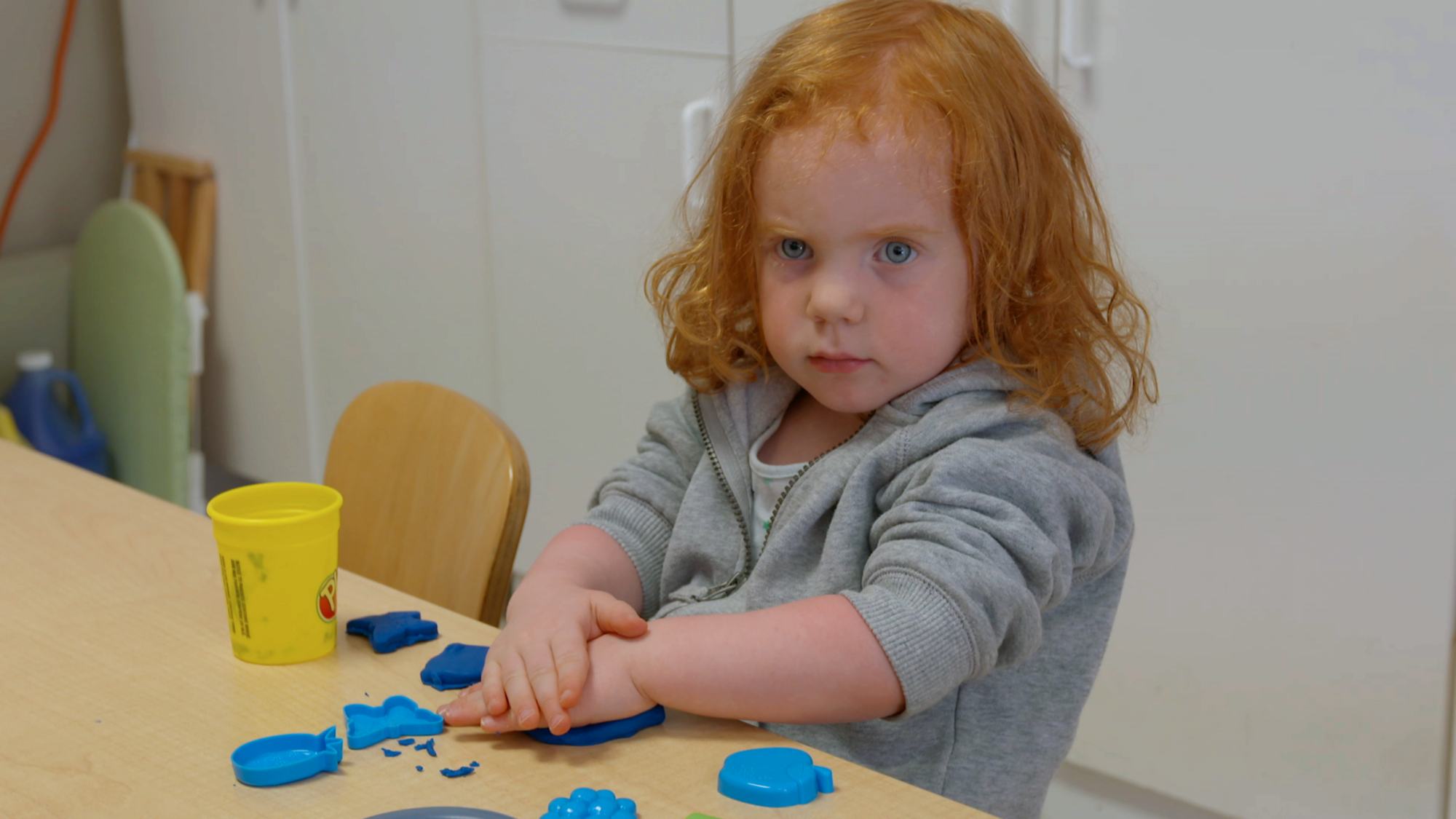 A young child with curly red hair playing with blue modeling clay at a table.