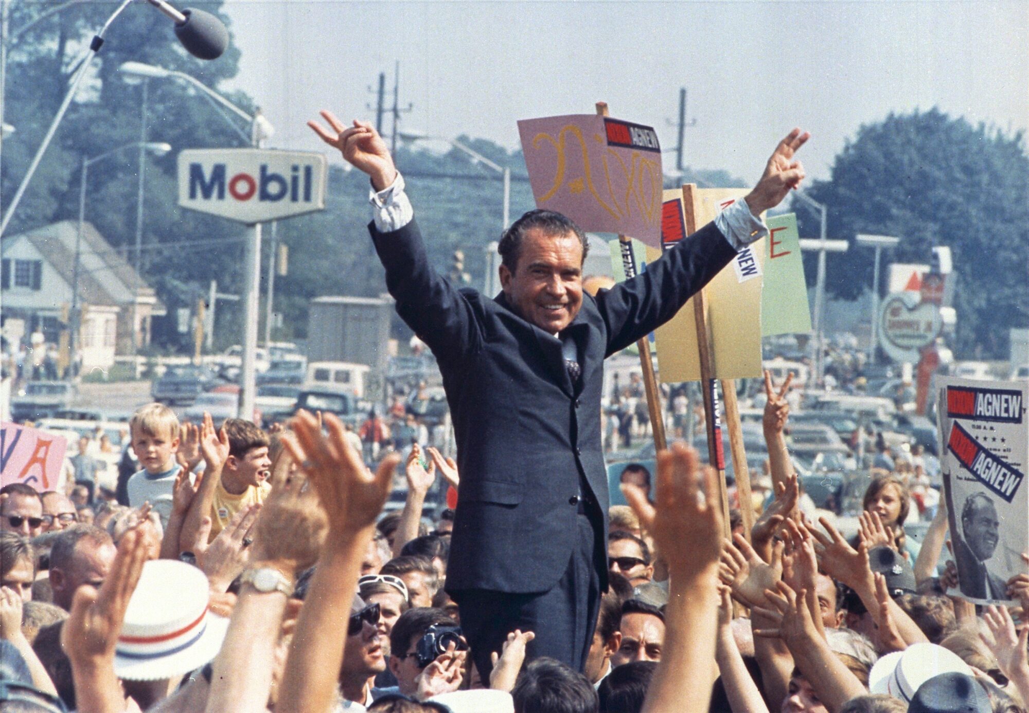 President Nixon in a dark suit gestures with a V-sign amid a cheering crowd, surrounded by signage and media equipment.