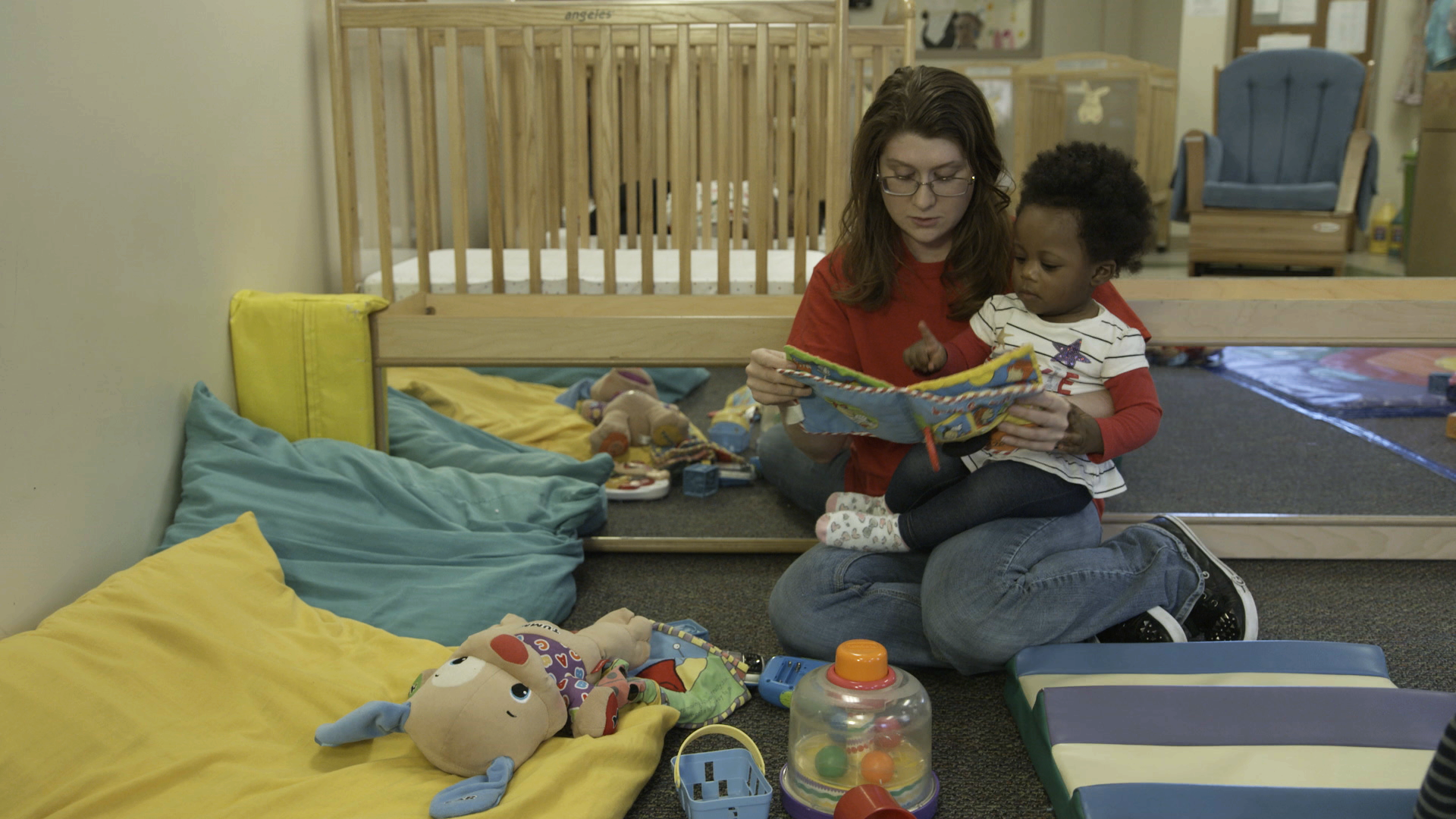 A woman reading to a child on her lap in a nursery setting with colorful cushions and toys around them.