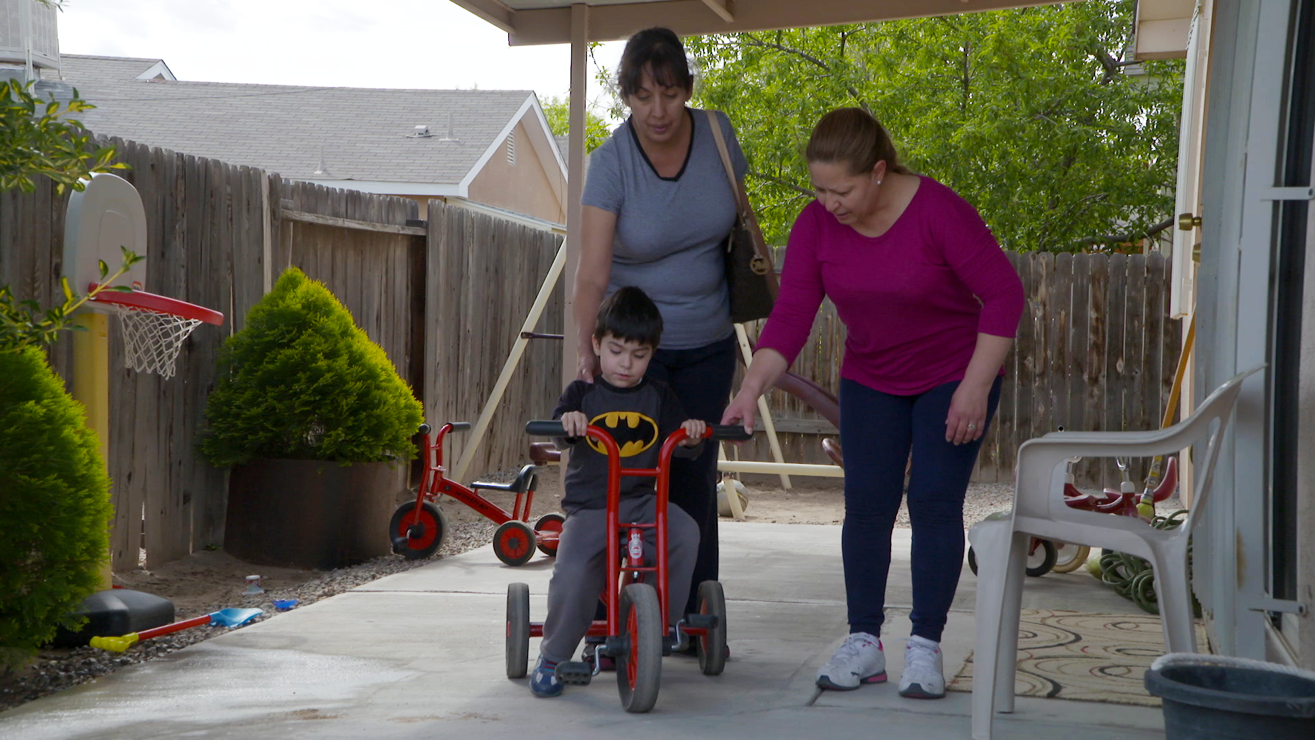 A child on a red tricycle with two adults assisting, on a pathway beside a wooden fence and plants.