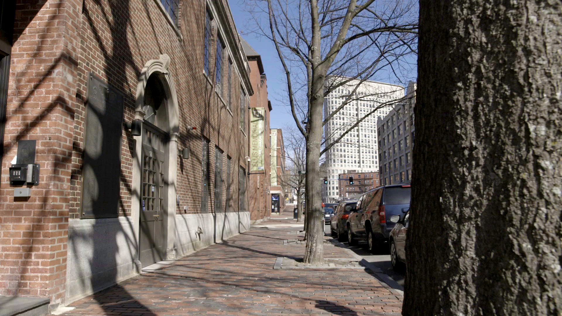 Brick sidewalk in an urban area with a historic building and modern skyscrapers in the background. Tree shadows stretch across the scene.