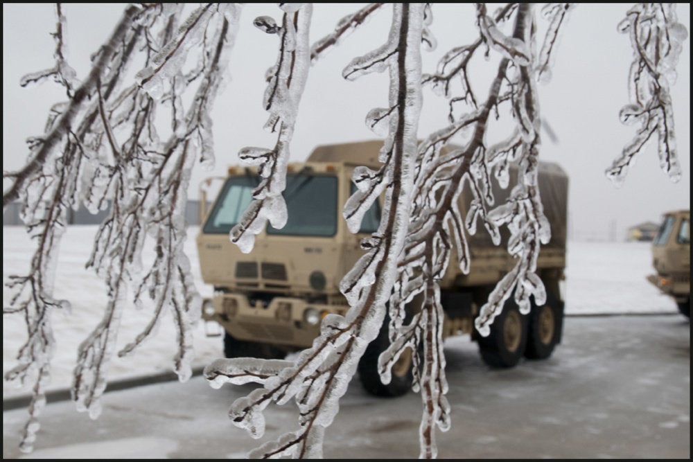 Ice-coated branches in the foreground with a military truck in the snowy background.