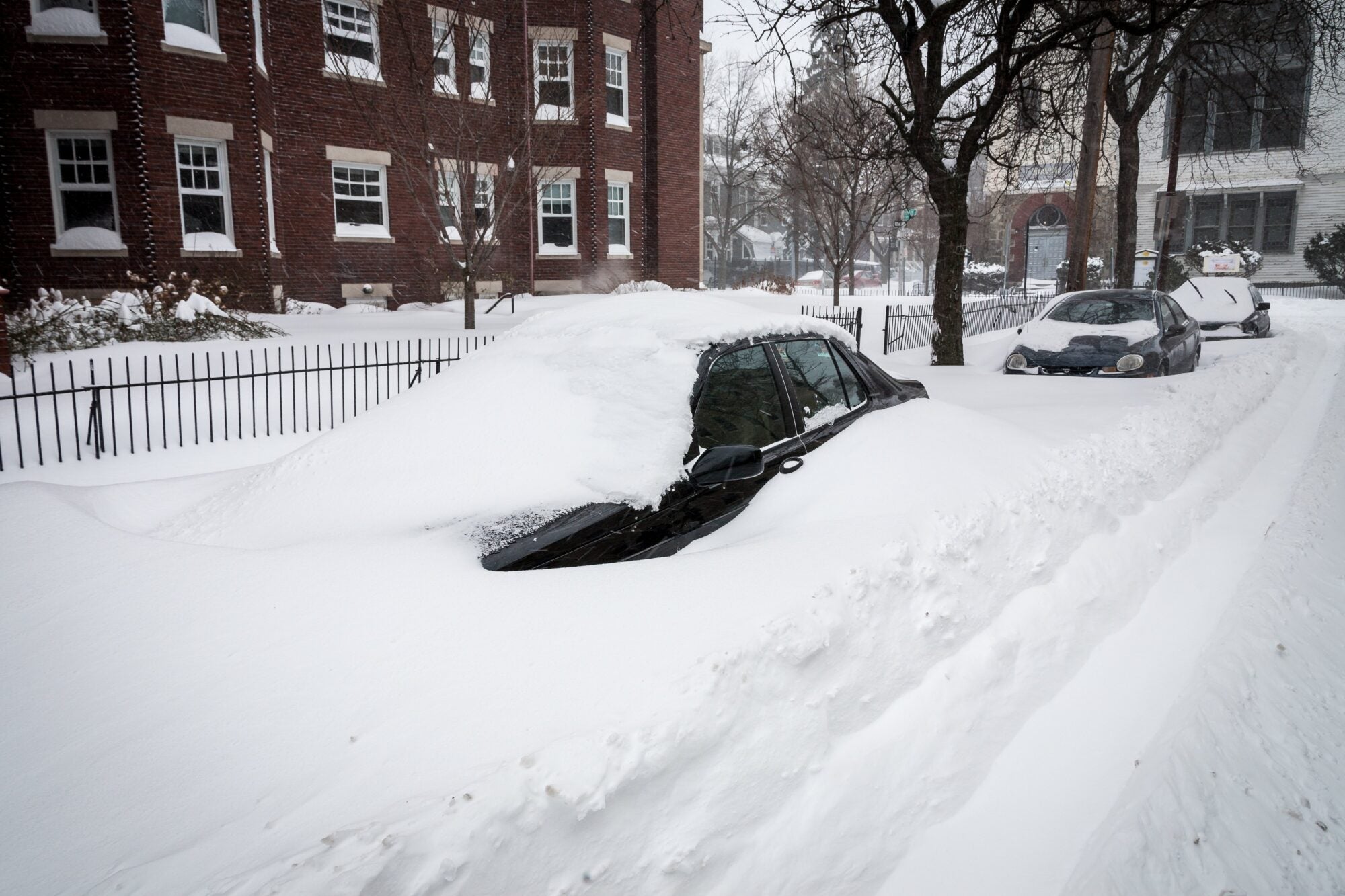 A black car buried under snow on a city street lined with brick buildings and trees.