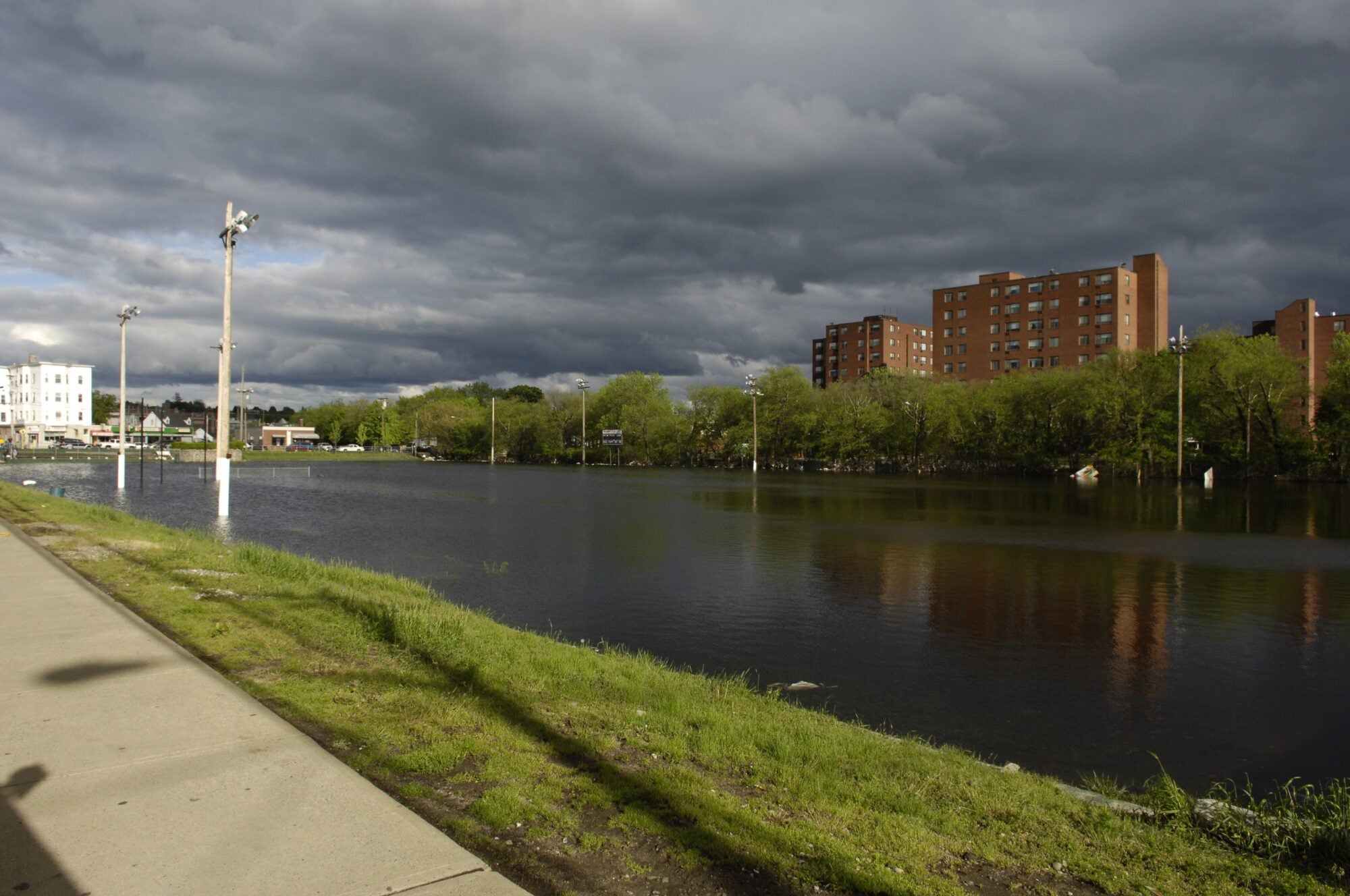 Flooded area with submerged lamp posts, bordered by a path and tall red brick buildings in the background under a stormy sky.