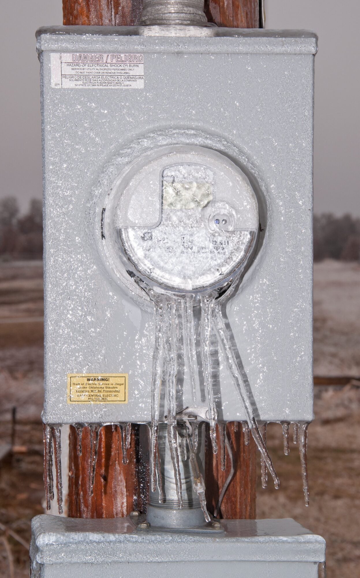 An electrical meter box covered in ice with hanging icicles. Two warning labels are visible.