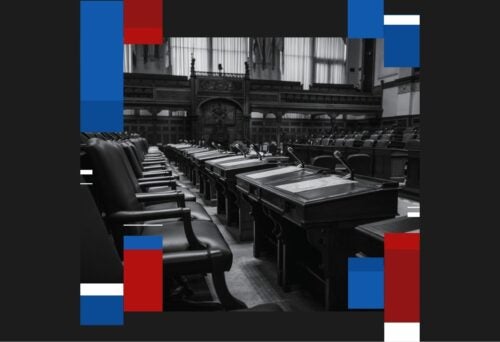 Black and white image of an empty legislative assembly room with leather chairs and wooden desks, framed by red, blue, and white graphics.