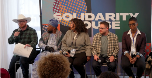 The image depicts a panel of five individuals seated on a stage in front of a colorful backdrop. The backdrop features abstract patterns in circles and rectangles with a prominent text stating 