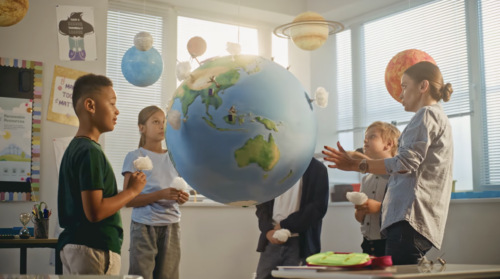 Three children and a teacher stand near a huge replica of the globe to understand its scale and relationship to planets.