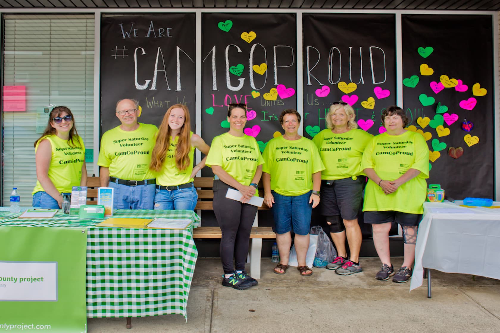 A group of seven people wearing bright green t-shirts standing in front of a black banner with colorful hearts and text.