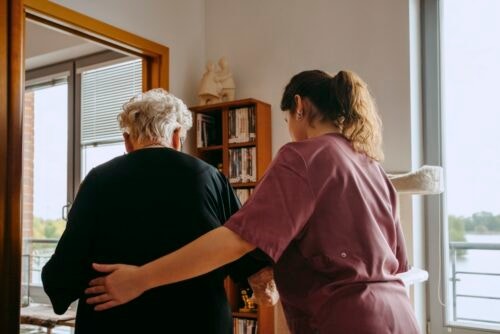 Rear view of female caregiver supporting senior woman while walking in apartment
