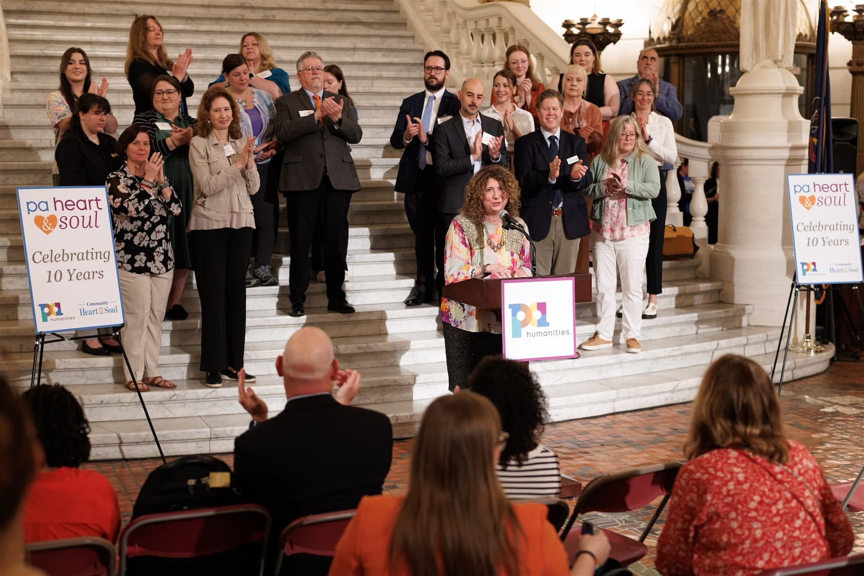 A group of people stands on a marble staircase with a speaker at a podium, celebrating an event.