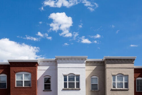 Row of four attached buildings with facades in red brick, white, and beige under a blue sky with clouds.