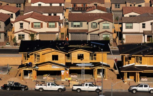 Workers construct large wooden house frames on a dry street in front of finished and painted similar homes.