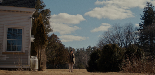 In a screencap from the film Sorry, Baby, someone stands in the forested yard of a rural New England home under a blue sky.