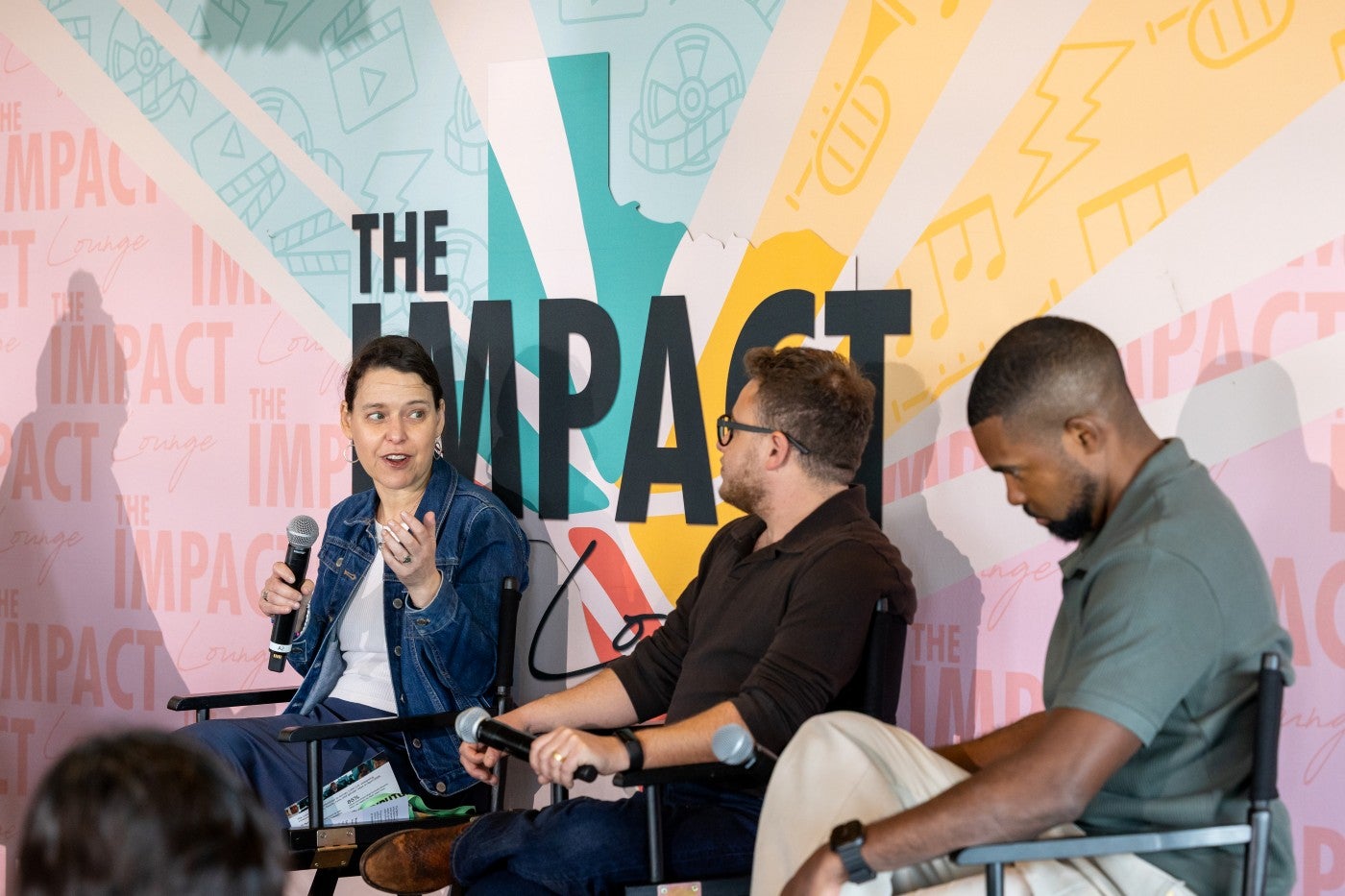 Vicki Shabo sits in a directors' chair in front of a pink, yellow, and teal banner, speaking into a microphone, next to Alex Schmider and Dennis St. Rose.