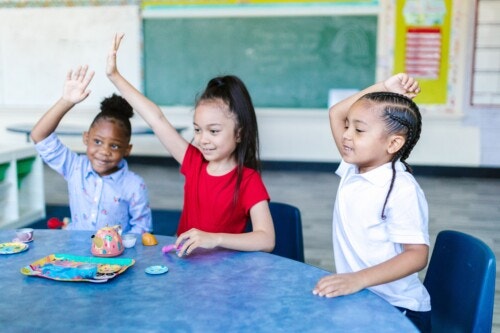 Three smiling young children sit at a classroom table, each raising one of their hands. Toys are spread across the table.