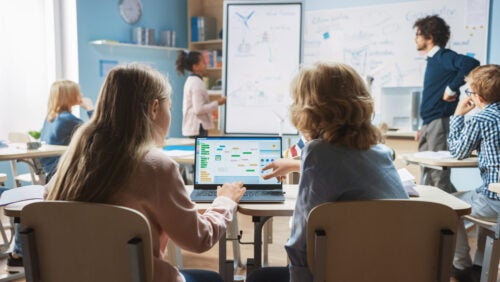 Two students on a computer and a teacher and student at a whiteboard