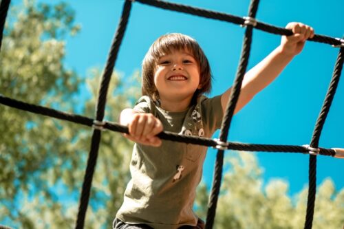 Photograph of smiling white boy with straight brown hair outside on a playground climbing a geodesic dome made of ropes