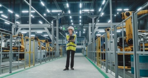 An engineer works on a laptop while walking through a factory with manufacturing robots.