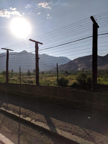 A barbed wire fence in the foreground with mountains and a bright sun in the background.