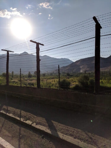 A barbed wire fence in the foreground with mountains and a bright sun in the background.