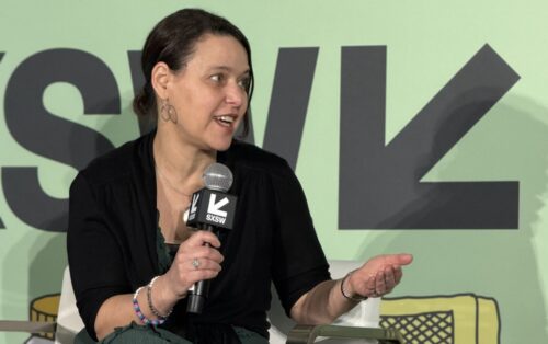 Vicki Shabo of New America's Entertainment Initiative is seated in front of a mint-green South by Southwest backdrop. She holds a branded handheld microphone as she speaks on a panel.