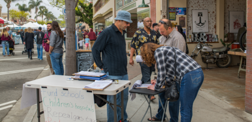 People gathered around a table at an outdoor market event, a woman writing on papers.