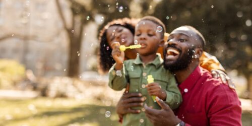Family blowing bubbles together outdoors in a sunlit park setting.