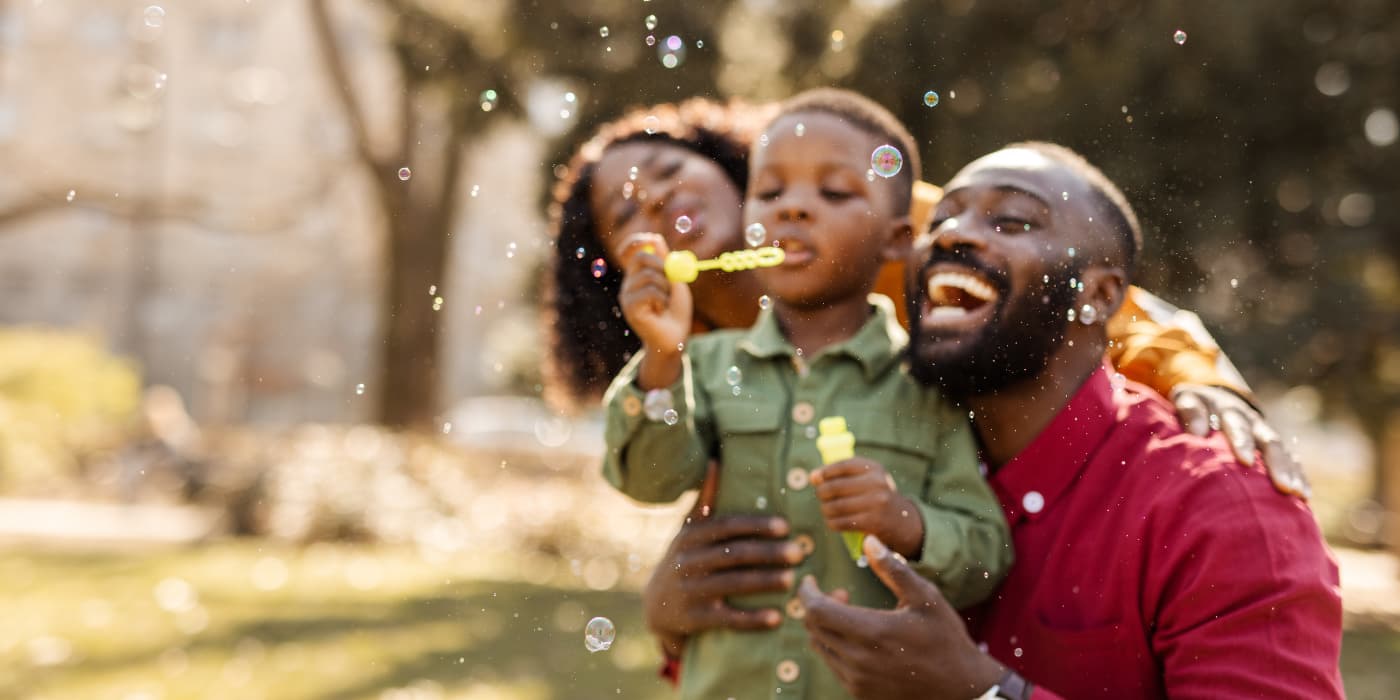 Family blowing bubbles together outdoors in a sunlit park setting.
