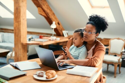 African-American girl using smart phone and sitting on mother's lap while she is working on laptop at home.