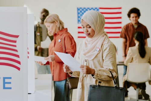 A woman in hijab reads a paper in front of a polling station as others around her also vote.