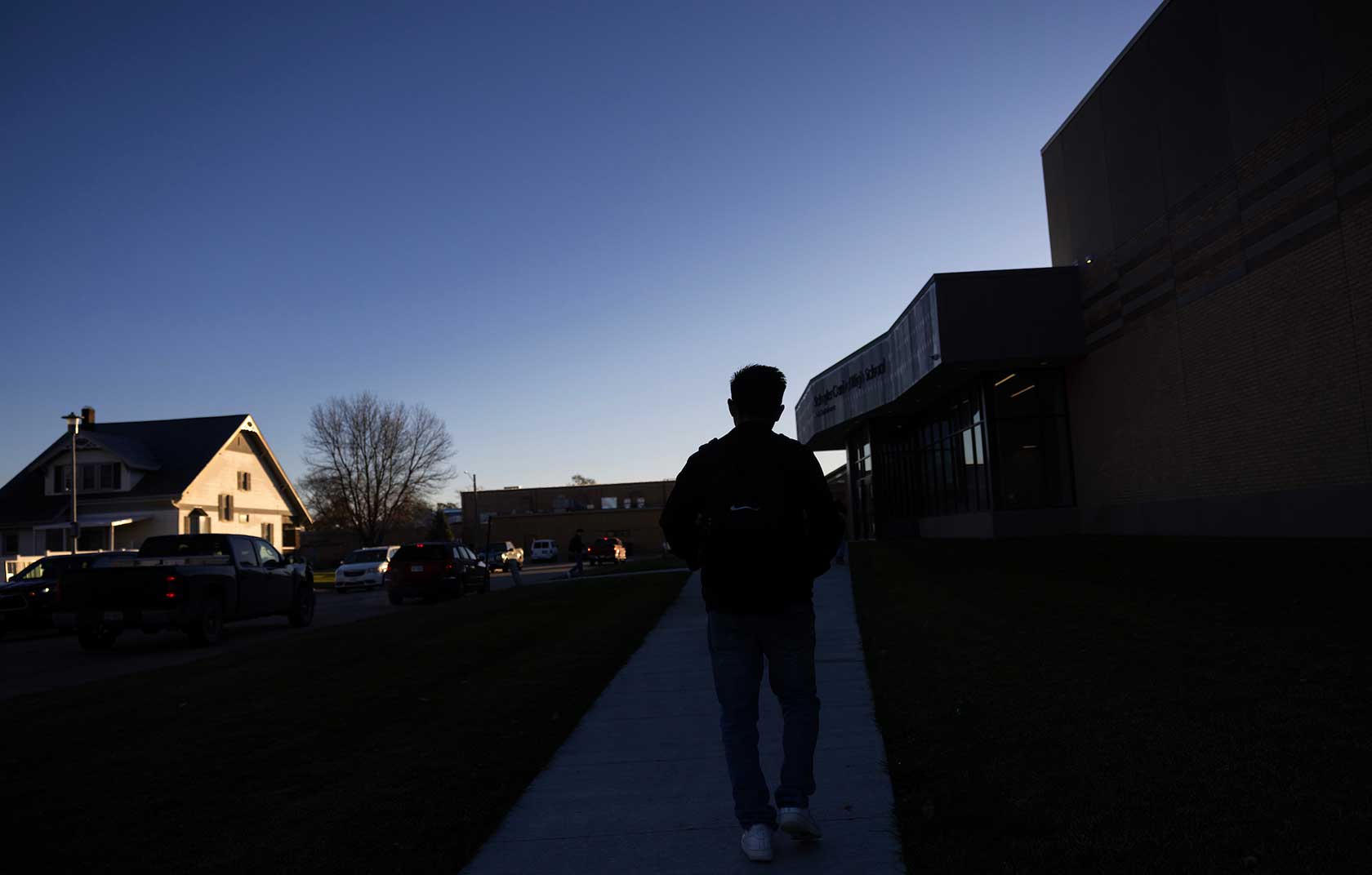 Silhouette of a person walking on a sidewalk near a modern school building at dusk, with a house and parked cars in the background.