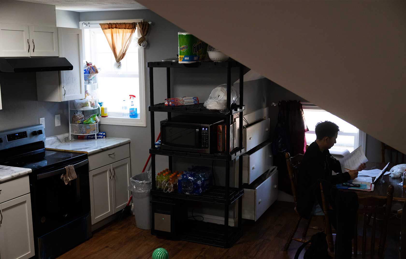 A cozy kitchen featuring white cabinets, a black stove, and a window with natural light. A person studies at a wooden table, surrounded by a black shelving unit filled with kitchen items and drinks.