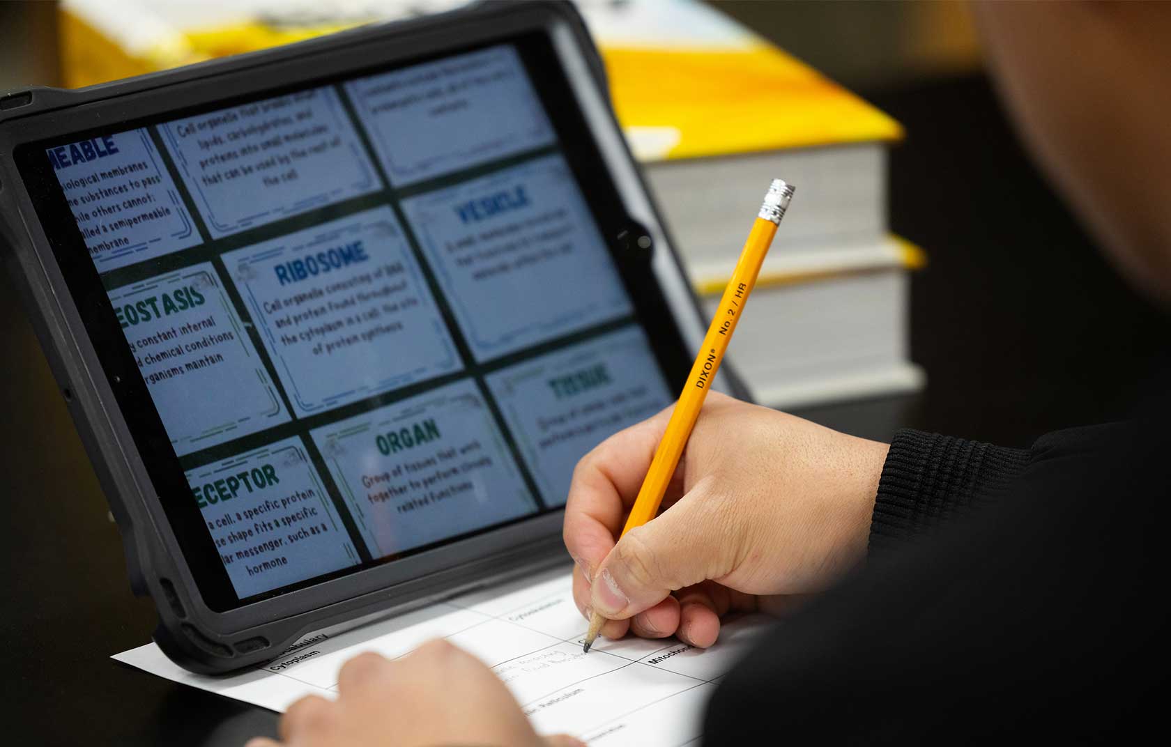 A person writes with a pencil on paper while engaged with a tablet displaying educational cards on biological terms, set against a dark surface.