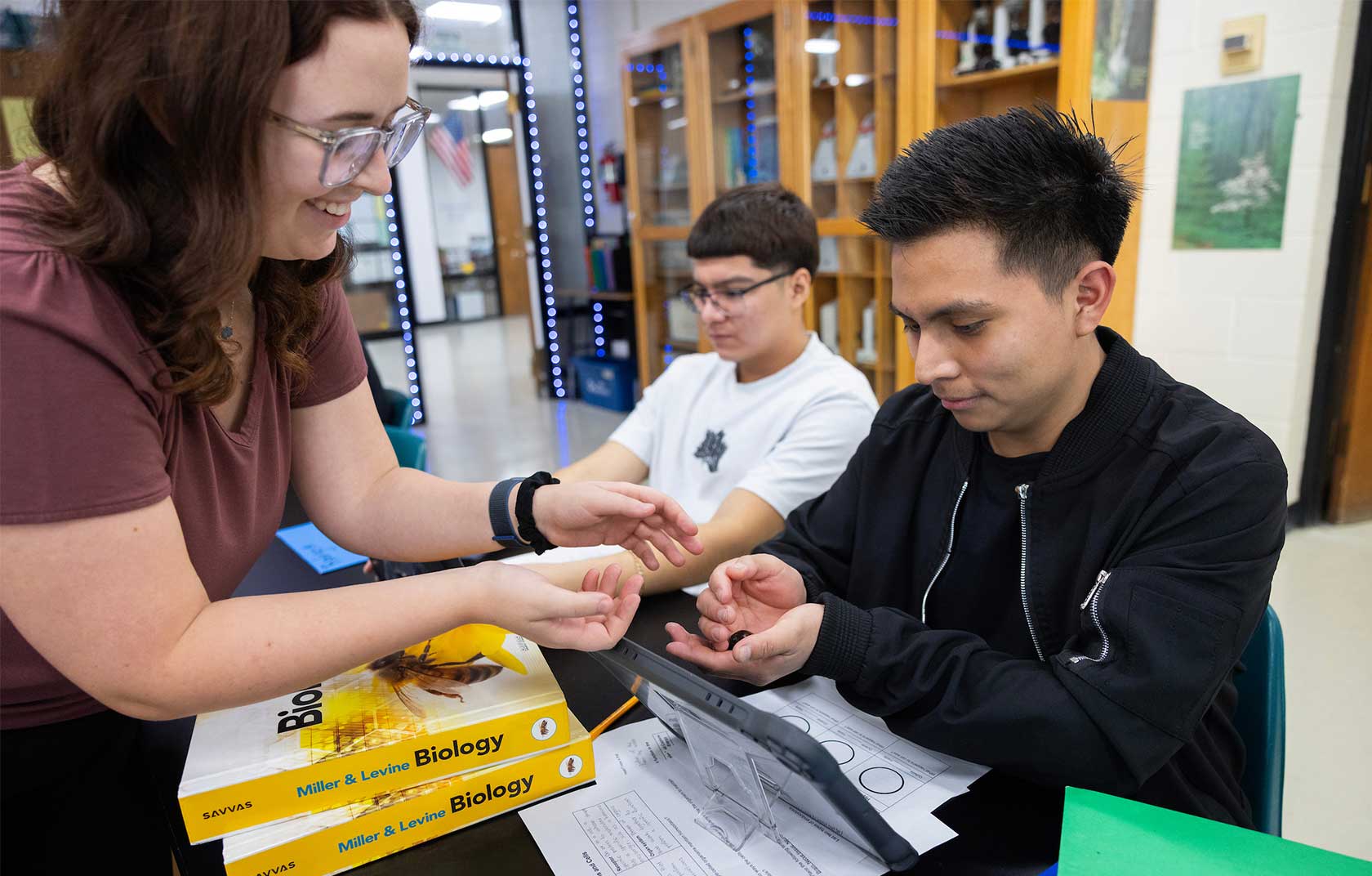 A female student assists a male student in examining an object in his hand during a biology class, while another male student observes. Biology textbooks and a tablet are on the table.