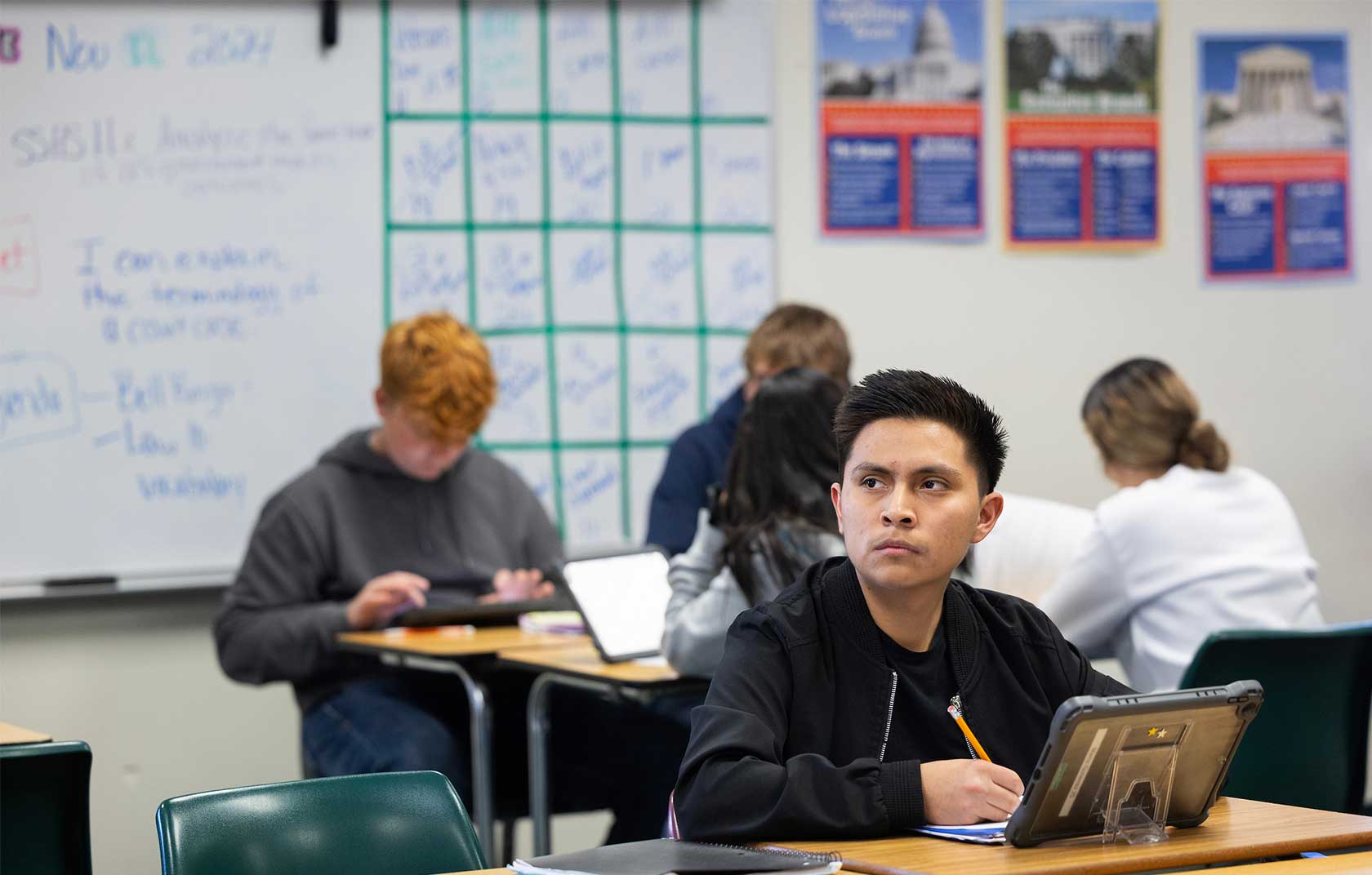 A focused student in a black jacket is writing with a pencil at a desk in a classroom. Other students are using tablets in the background, with a whiteboard filled with notes and calendar grids visible.